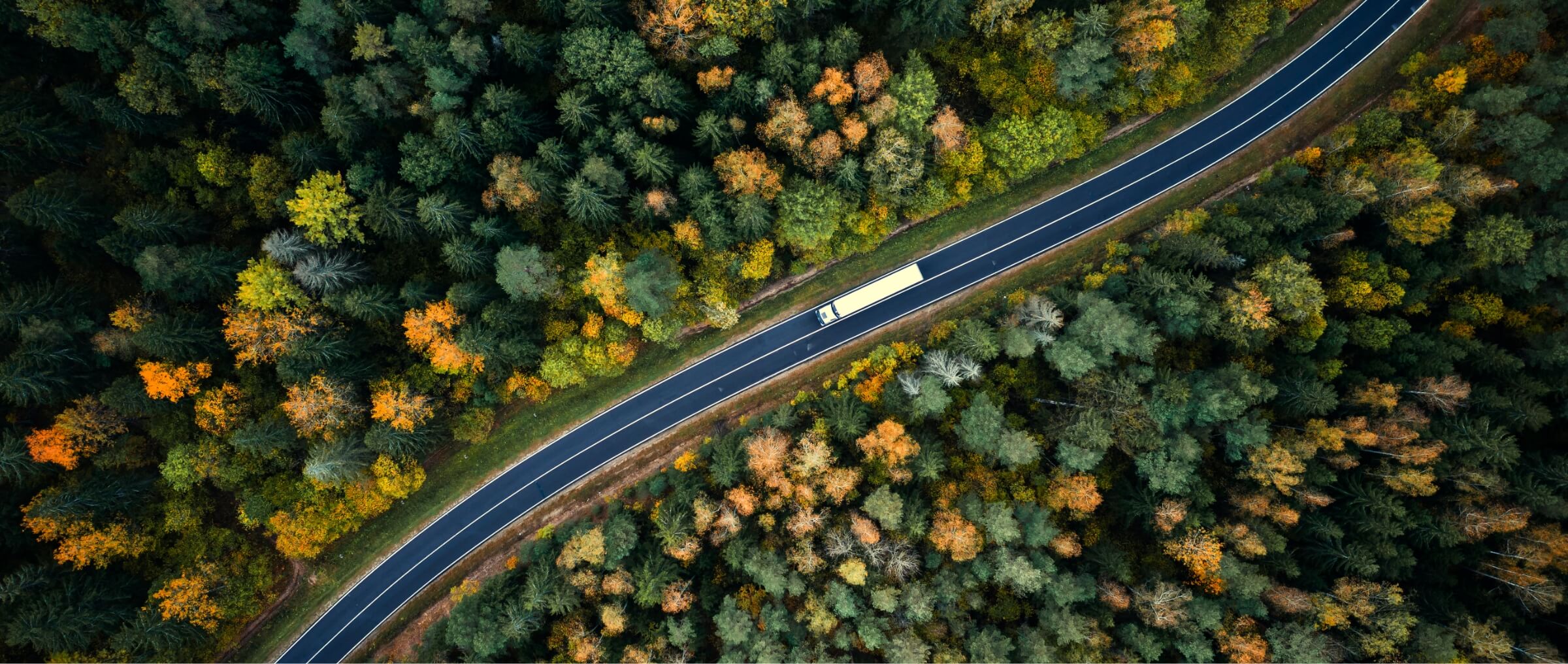 Aerial of a white transport truck driving on a highway surrounded with forest in the fall.