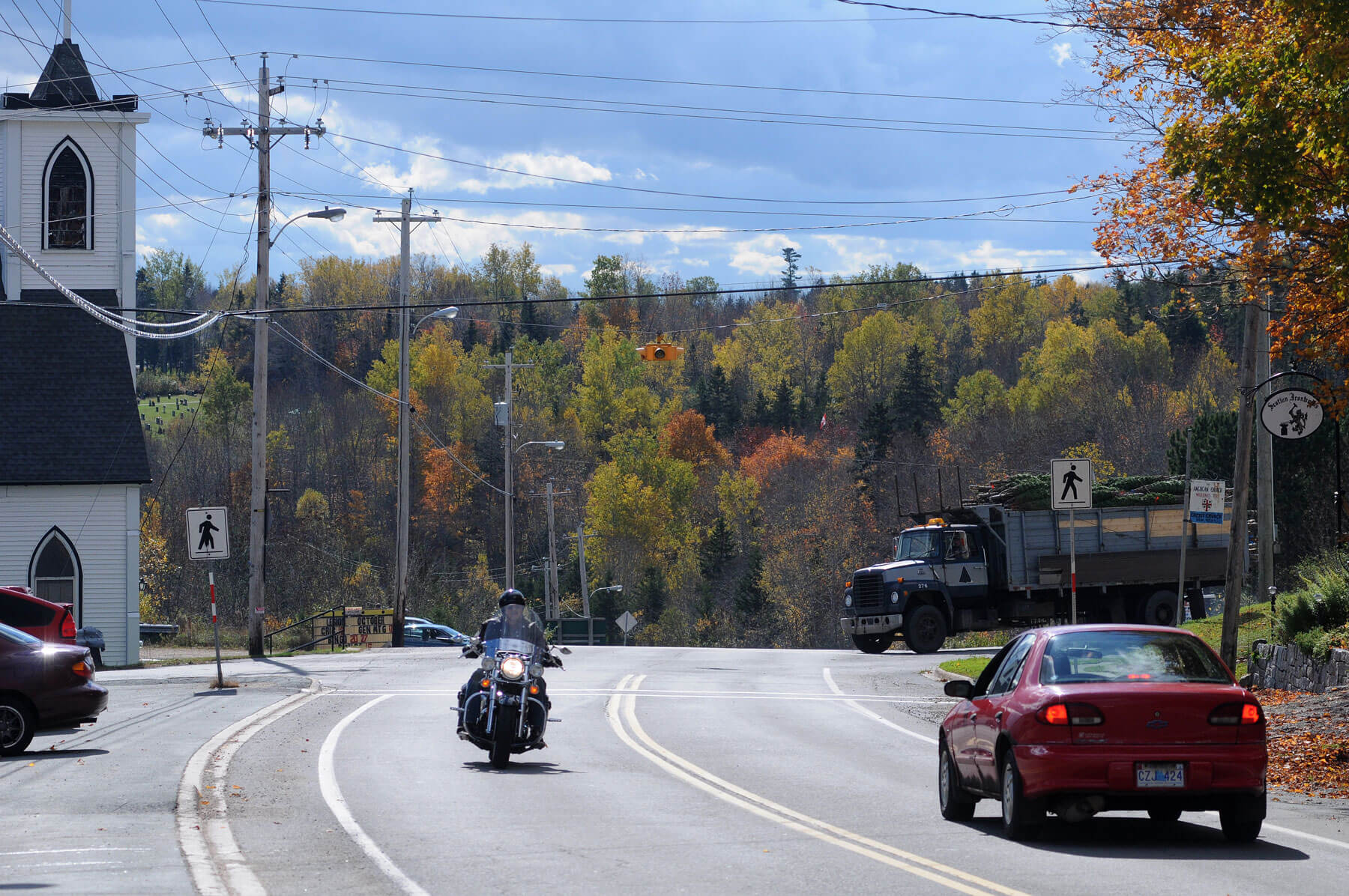 An intersection in New Ross. A motorcyclist travels toward the camera, a car travels away from the camera, and a truck carrying Christmas trees waits at a stop sign.