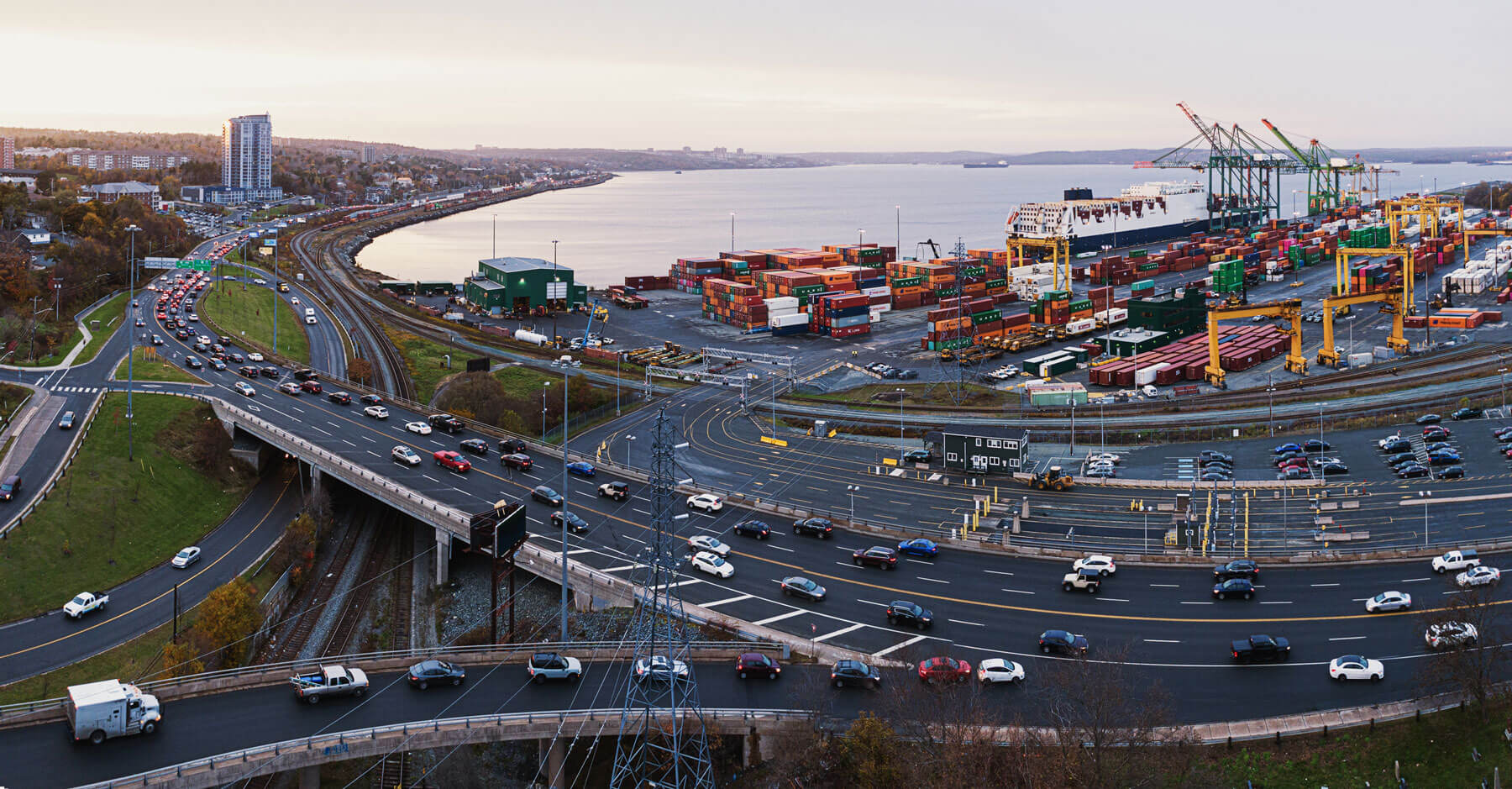 An aerial of Bedford Highway near the Windsor Street exchange. Cars and trucks travel inbound and outbound. The container pier and railway, along with the Bedford Basin, are seen in the background.