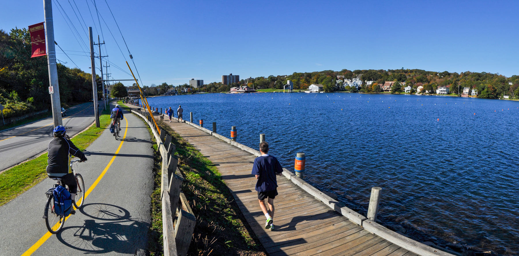 Two people cycle on the multi-use pathway next to Lake Banook in fall. A person runs next to them on the boardwalk.