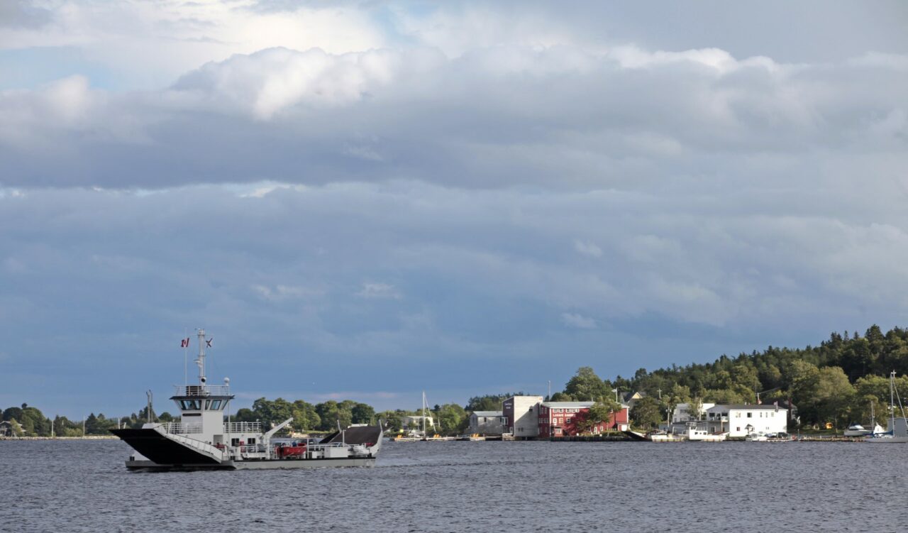 The LaHave ferry crosses the LaHave river against a cloudy sky. The Lahave Bakery is visible in the backgroumd.